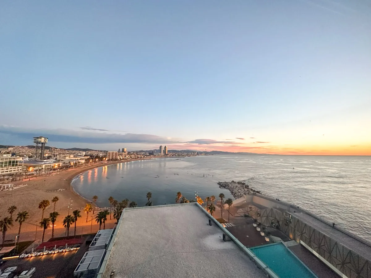 Panoramic view of Barcelona's coastline at dusk, featuring the sandy beach, a calm sea, and a vibrant sunset skyline.