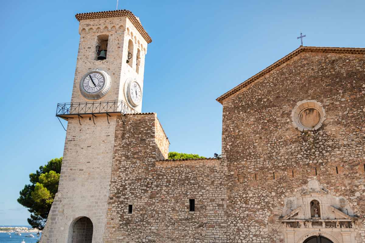 The historic clock tower of the Notre-Dame d'Esprance church standing tall against a clear blue sky in the old quarter of Cannes, with the fortified walls of the medieval building visible.