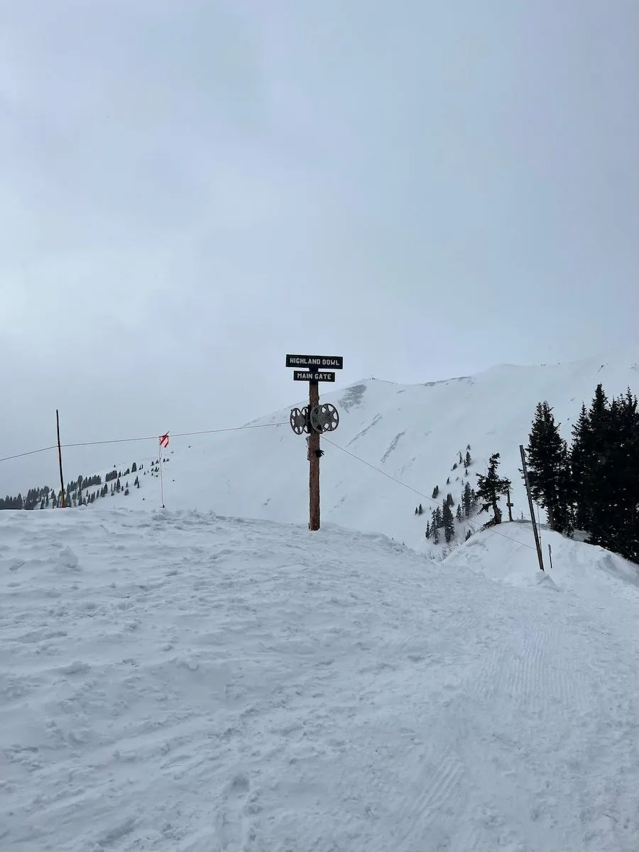 A signpost at the top of a snowy Aspen mountain with the text "Highland Bowl" indicating ski routes under an overcast sky.