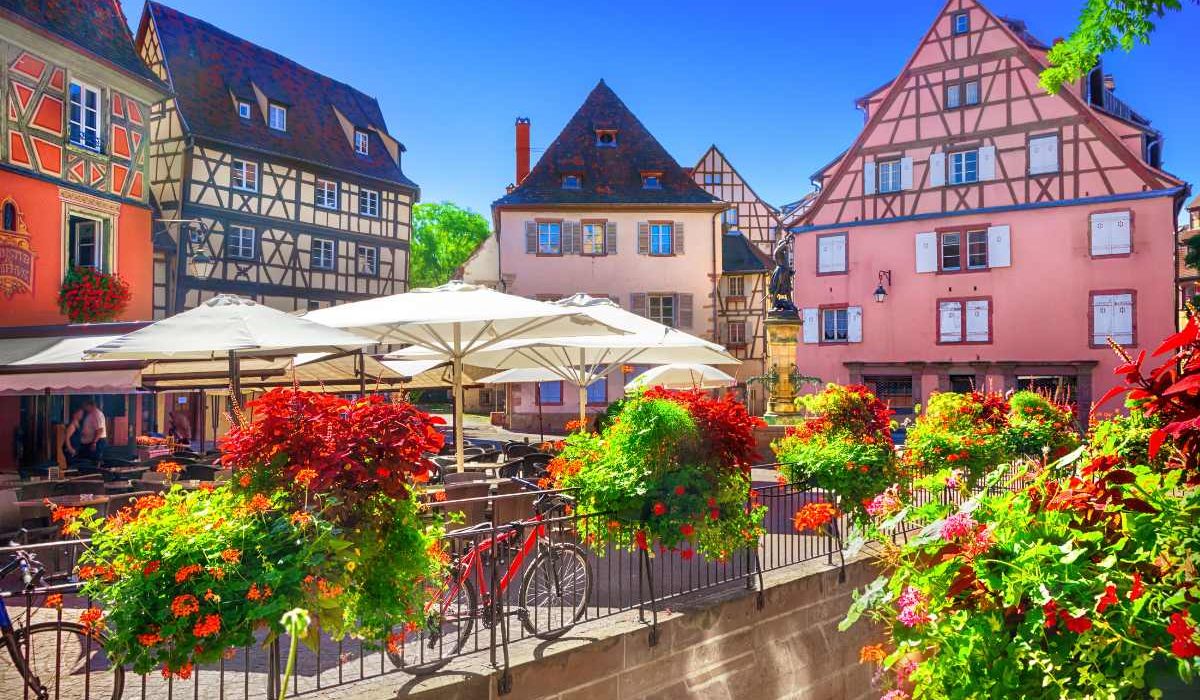 A bright, sunny day in an Alsace village, where colorful half-timbered buildings frame a lively square with outdoor caf umbrellas, vibrant flowers, and a bicycle leaning against a railing, inviting a leisurely day out.