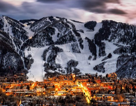 A twilight view of Aspen, with warm city lights illuminating the town against the snowy backdrop of ski runs on the mountains. This photo perfectly sums up why Aspen is worth visiting.