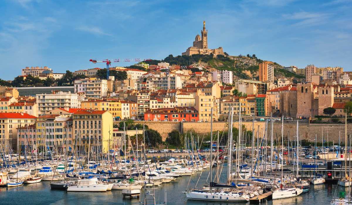 he bustling Marseille Old Port filled with boats and yachts, surrounded by buildings with the Notre Dame de la Garde basilica sitting on a hill in the background."
