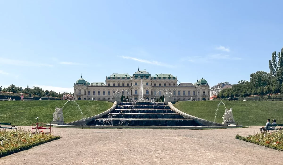 A grand fountain in the foreground with water arching into the air, set against the backdrop of a regal, historical palace surrounded by manicured lawns. Summer is the best time to visit vienna, as you can see the great weather in this picture