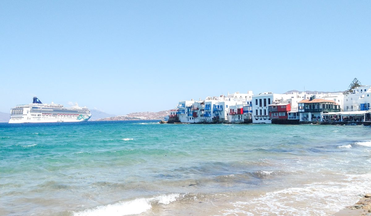 A stunning seascape featuring the famed Little Venice of Mykonos, where colorful buildings perch precariously on the water's edge, with waves lapping at the foundations, under a sky with cotton-like clouds.