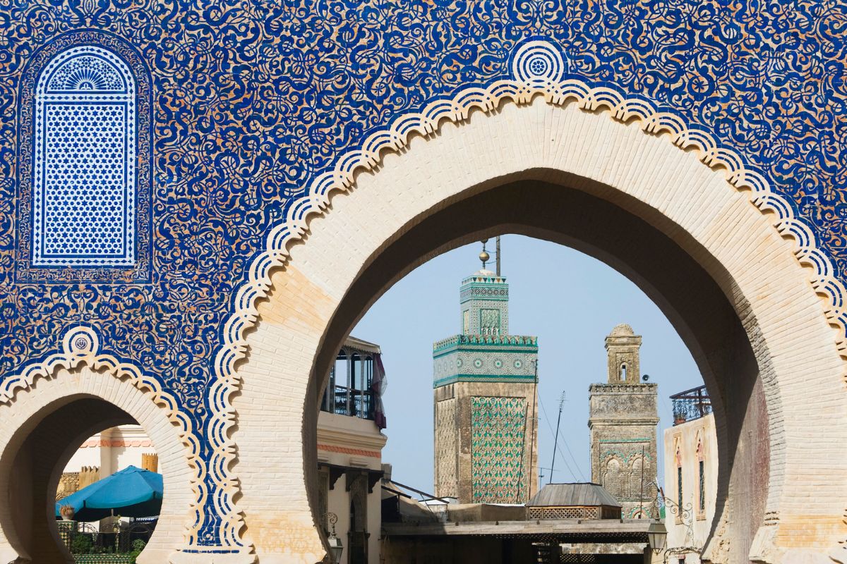 A detailed view of Moroccan architecture with intricate blue and white ceramic tiles adorning an archway, framing a distant minaret against a clear sky in Fes
