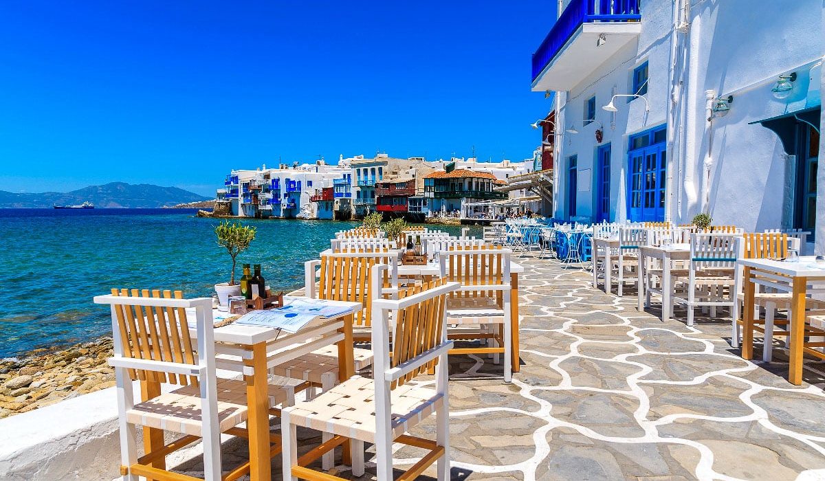 A bright, sunny view of a coastal cafe in Mykonos. White chairs and tables are arranged on a stone-paved terrace, with the tranquil sea stretching out in the background, and traditional whitewashed buildings with blue accents line the waterfront.