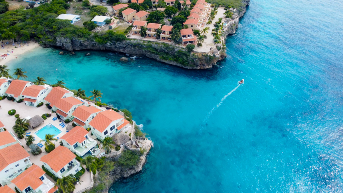 Aerial view of Playa Lagun, Curacao showing a bustling beach nestled between rugged cliffs and turquoise waters. A small boat is drifting in the foreground