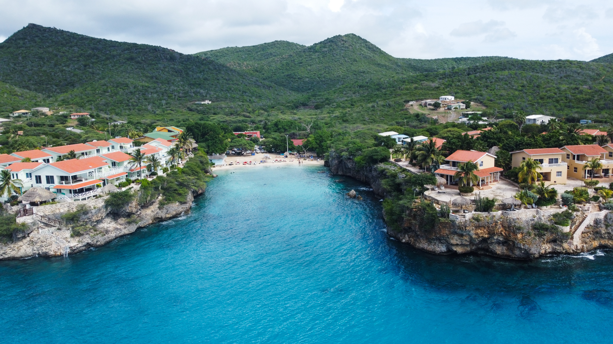 Aerial view of Playa Lagun, Curacao showing a bustling beach nestled between rugged cliffs and turquoise waters