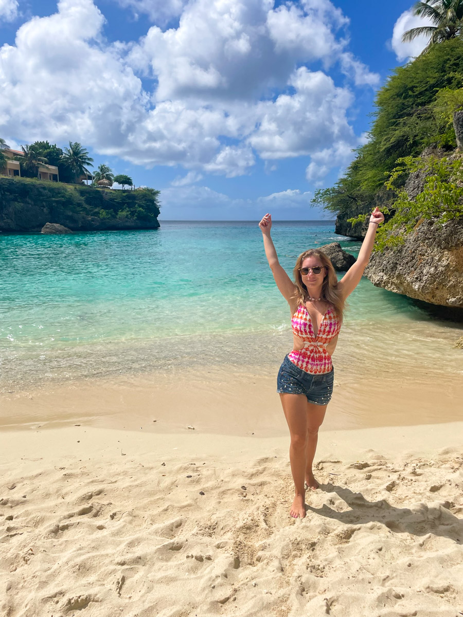 "The author's wife celebrating with raised arms on the sandy beach of Playa Lagun, Curacao, with tropical waters and cliffside villas in the background."