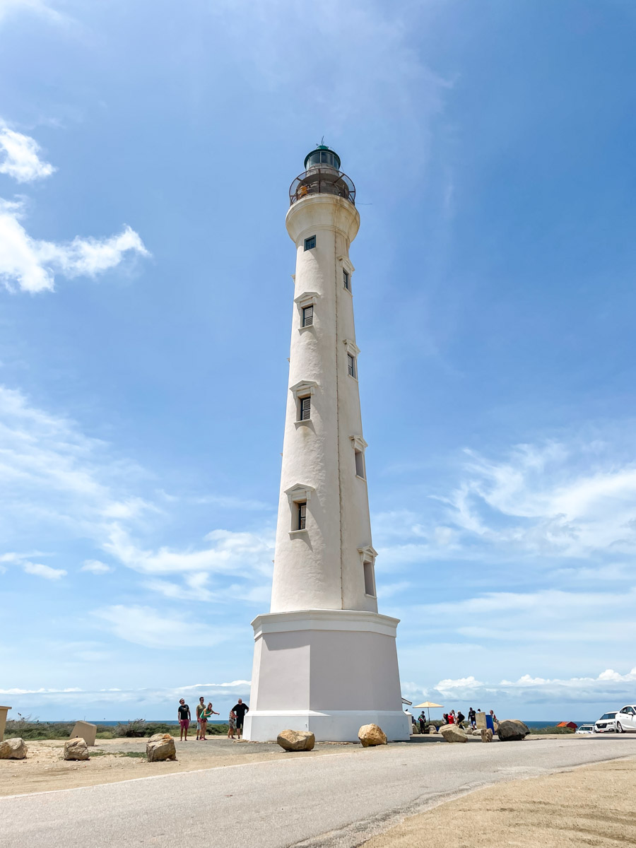 White historical lighthouse against a bright blue sky with people around its base on a sunny day.