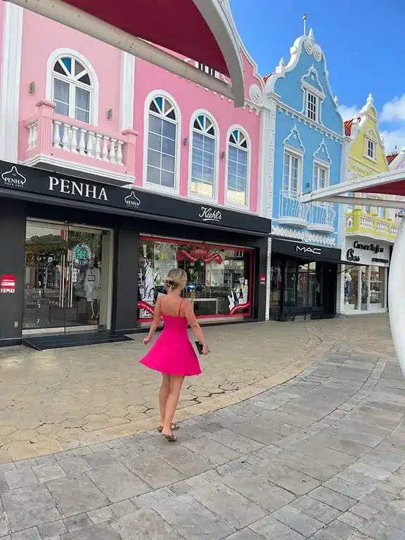 Picture of the author's wife going shopping in a pink dress in Aruba. There are cute, colorful houses behind her. 