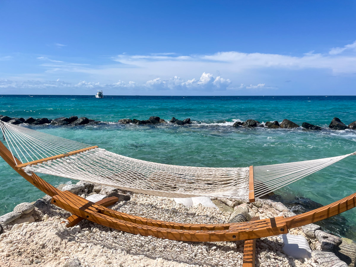 Relaxing hammock set up on a rocky beach with a vibrant blue ocean and a yacht in the background. Renaissance island is one of the most expensive day trips in Aruba on vacation.
