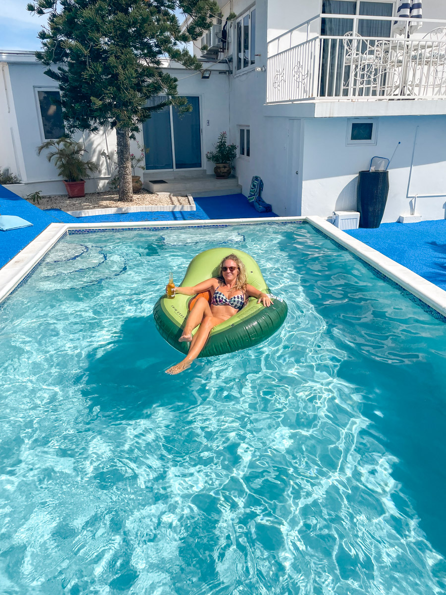 Woman lounging in a green inflatable ring, enjoying a drink in a sunny pool area with a white building background. Renting a house can be a good way to avoid the expensive hotel costs in Aruba.